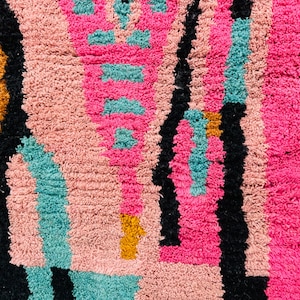 May include: Close-up of a colorful, textured rug with a geometric pattern. The rug features a combination of pink, black, teal, and gold colors. The design includes vertical and horizontal lines and shapes.