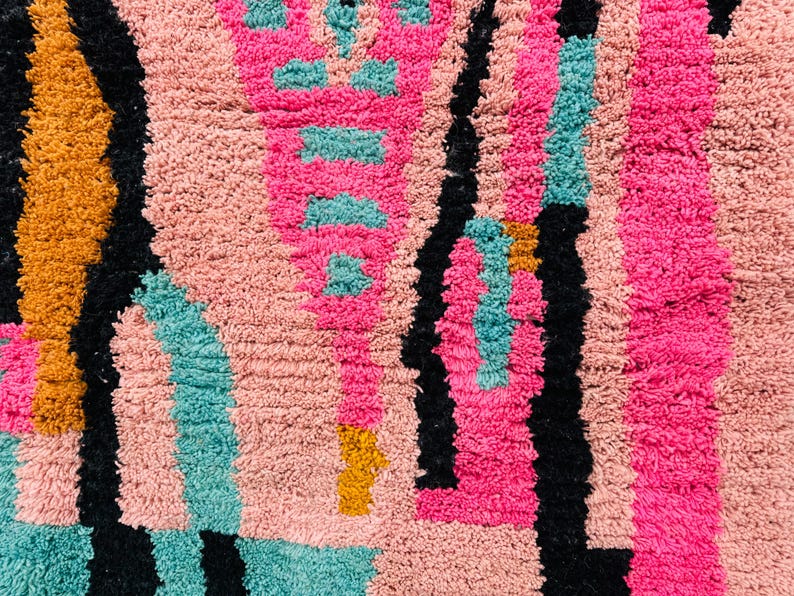 May include: Close-up of a colorful, textured rug with a geometric pattern. The rug features a combination of pink, black, teal, and gold colors. The design includes vertical and horizontal lines and shapes.