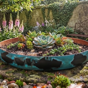 May include: A shallow, round metal planter with a turquoise and black patina finish, filled with a variety of green and reddish succulents planted in dark soil, featuring a copper-coloured interior rim and a wide, low-profile circular design.