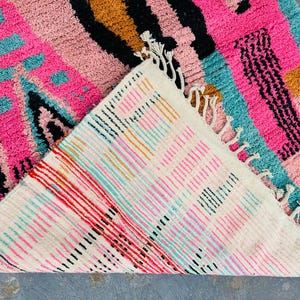 May include: A vibrant, colorful rug with a geometric design. The rug features a mix of pink, teal, black, and gold hues. The underside of the rug is visible, showcasing a striped pattern with red, pink, and black lines. The rug has a textured surface and fringe.