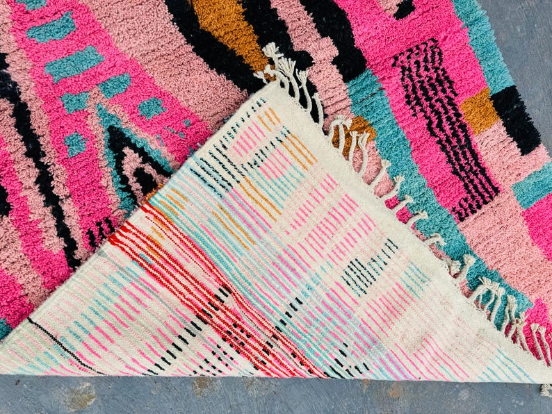 May include: A vibrant, colorful rug with a geometric design. The rug features a mix of pink, teal, black, and gold hues. The underside of the rug is visible, showcasing a striped pattern with red, pink, and black lines. The rug has a textured surface and fringe.