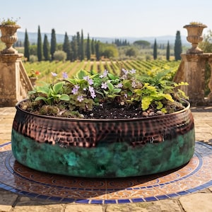 May include: A large, round, hammered copper planter with a vibrant teal patina finish on the lower half. The planter is filled with dark soil, green foliage, and small purple flowers, sitting on a circular tiled patio surface.