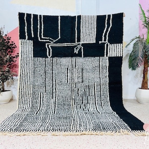 Puede incluir: Alfombra con estampado geométrico en blanco y negro con flecos. La alfombra presenta una combinación de rayas verticales y formas abstractas. La alfombra está hecha de un material grueso y tejido, y se muestra sobre un fondo neutro.