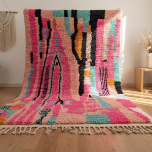 May include: A colorful, abstract rug with a pink, teal, black, and orange design. The rug has a textured appearance and a fringe along the bottom edge. The rug is displayed on a wooden floor.