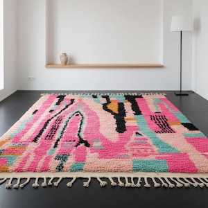 May include: A rectangular rug with a vibrant abstract design. The rug features a mix of pink, black, teal, and yellow shapes. It has a cream-colored fringe along the edges. The rug is placed on a dark floor.