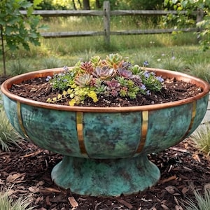 May include: A large, round, pedestal-style planter with a weathered teal and green patina finish, accented by vertical polished brass strips and a copper-toned rim, filled with a variety of small, colourful succulent plants in dark soil.