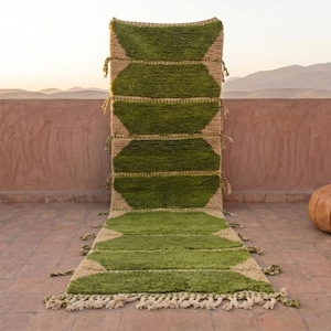 May include: A green and beige geometric rug with a fringe, displayed outdoors. The rug has a series of green diamond shapes on a beige background. A brown leather ottoman is to the right of the rug. Mountains are in the background.