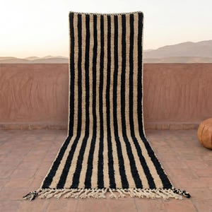 May include: A black and beige striped rug with a fringed edge is displayed outdoors. A round, brown leather ottoman sits to the right of the rug. The background features a terracotta-colored wall and a mountain range.