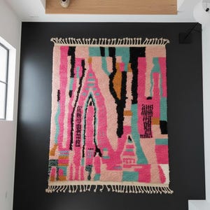 May include: A rectangular, abstract rug with a vibrant design. The rug features a mix of pink, black, teal, and gold vertical shapes against a light background. It has a fringed edge and is hanging on a black wall.
