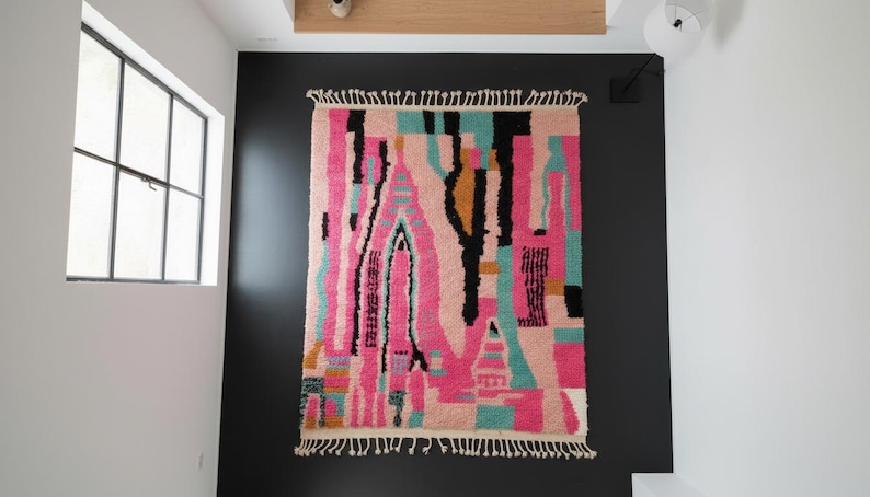 May include: A rectangular, abstract rug with a vibrant design. The rug features a mix of pink, black, teal, and gold vertical shapes against a light background. It has a fringed edge and is hanging on a black wall.