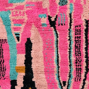 May include: Close-up of a vibrant, textured rug with a geometric design. The rug features a mix of bright pink, teal, black, and tan colors. The pattern includes vertical and horizontal lines, creating a modern, abstract look. The rug's texture is thick and plush.