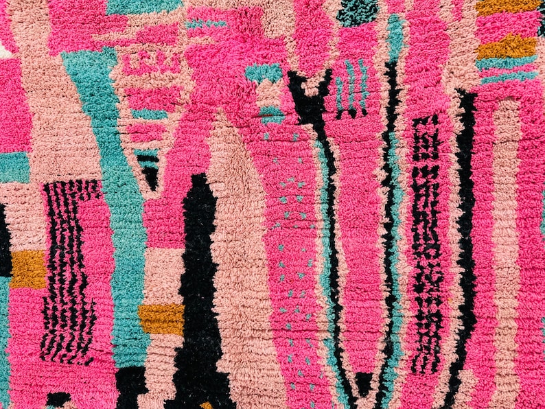 May include: Close-up of a vibrant, textured rug with a geometric design. The rug features a mix of bright pink, teal, black, and tan colors. The pattern includes vertical and horizontal lines, creating a modern, abstract look. The rug's texture is thick and plush.