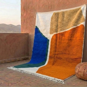 May include: A colorful, geometric rug with a white, blue, tan, and orange design. The rug is leaning against a terracotta wall. A brown leather ottoman sits on the floor next to the rug. The background shows a mountain range.
