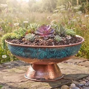 May include: A round, pedestal-style planter bowl featuring a hammered copper base and a textured, teal-patina finish on the upper exterior. The bowl is filled with dark soil and a variety of green and purple succulent plants of different shapes.