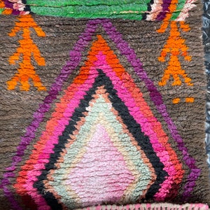 May include: A colorful Moroccan rug with a diamond pattern in shades of pink, orange, purple, and green. The rug has a brown background and is made of wool.