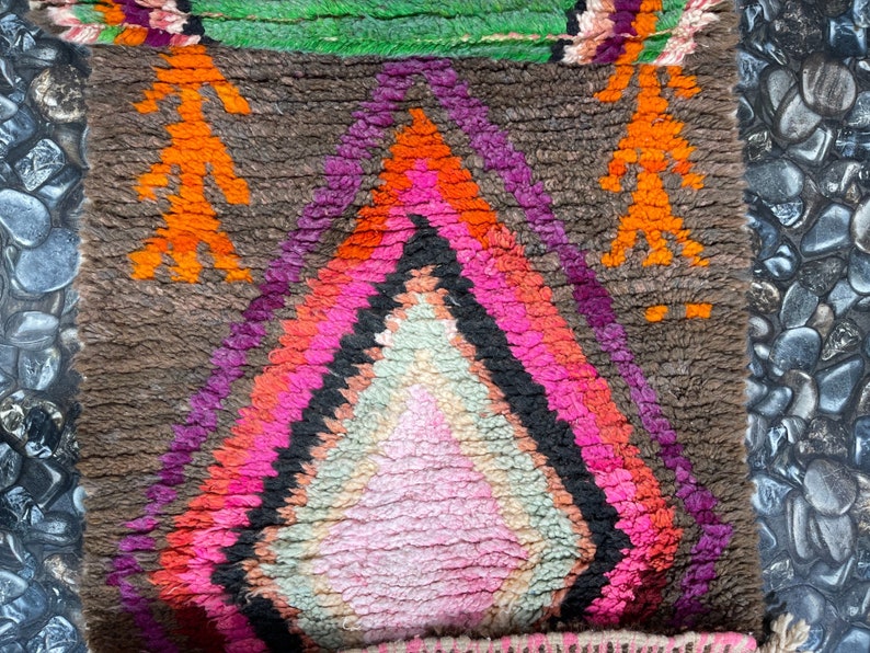 May include: A colorful Moroccan rug with a diamond pattern in shades of pink, orange, purple, and green. The rug has a brown background and is made of wool.