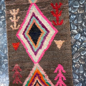 May include: A brown rug with a diamond pattern in pink, white, green, orange, and purple. The rug has a fringe on the top and bottom.