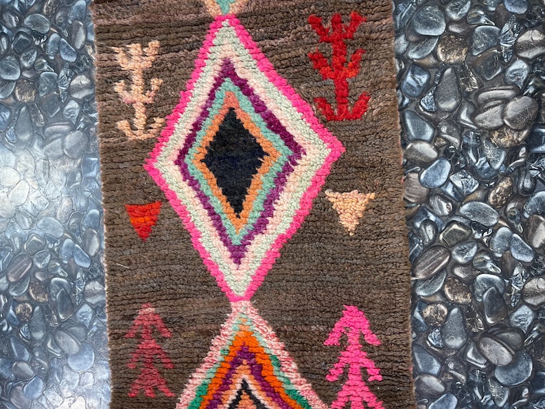 May include: A brown rug with a diamond pattern in pink, white, green, orange, and purple. The rug has a fringe on the top and bottom.