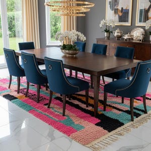 May include: A dining room set featuring a dark wood table surrounded by eight blue velvet chairs. A colorful rug with pink, turquoise, and black patterns sits beneath the table. The room has neutral-colored curtains and a chandelier.