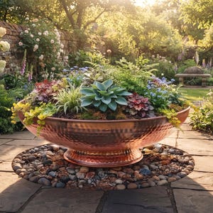 May include: A large, round, hammered copper bowl planter with a pedestal base, filled with a variety of green and reddish succulents and small foliage plants, resting on a circular bed of smooth, multi-colored river stones on a stone patio.