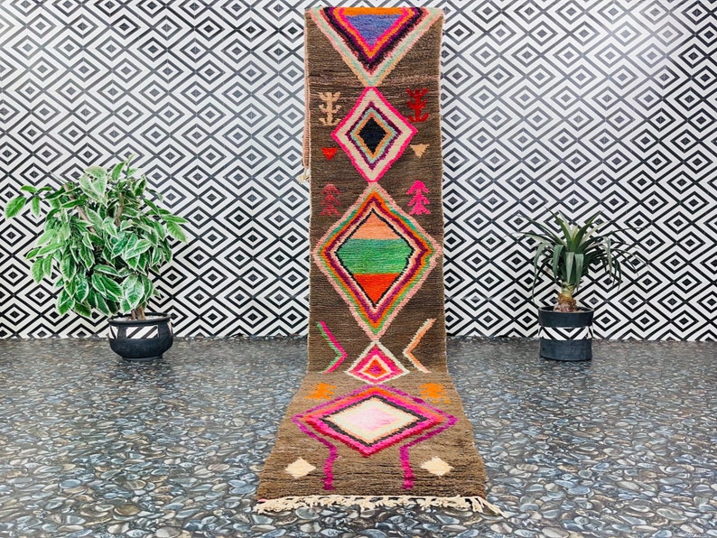 May include: A long, narrow rug with a brown background and colorful geometric patterns in shades of pink, orange, green, and blue. The rug has a fringe on both ends.