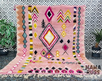 May include: Pink Moroccan rug with a geometric pattern in shades of pink, yellow, green, blue, and black. The rug has a fringe border and is made of wool.
