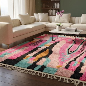May include: A colorful area rug with a geometric design. The rug features a mix of pink, black, teal, and yellow rectangular shapes. It has a fringed edge and is placed on a hardwood floor in a living room setting.