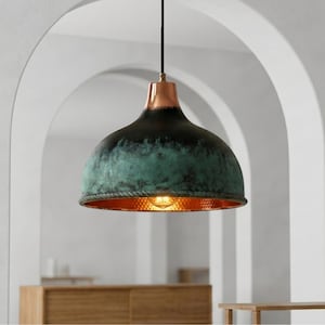 Op de afbeelding: Een koepelvormige hanglamp met een verweerde groenblauwe en zwarte patina-afwerking aan de buitenkant. Het bovenste gedeelte is van gepolijst koper en de binnenkant heeft een getextureerde, gehamerde koperen afwerking met een brandende gloeilamp.