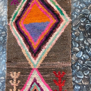 May include: A brown rug with a colorful diamond pattern in the center. The diamond is made up of pink, orange, purple, and green stripes. The rug has a fringe on the bottom.