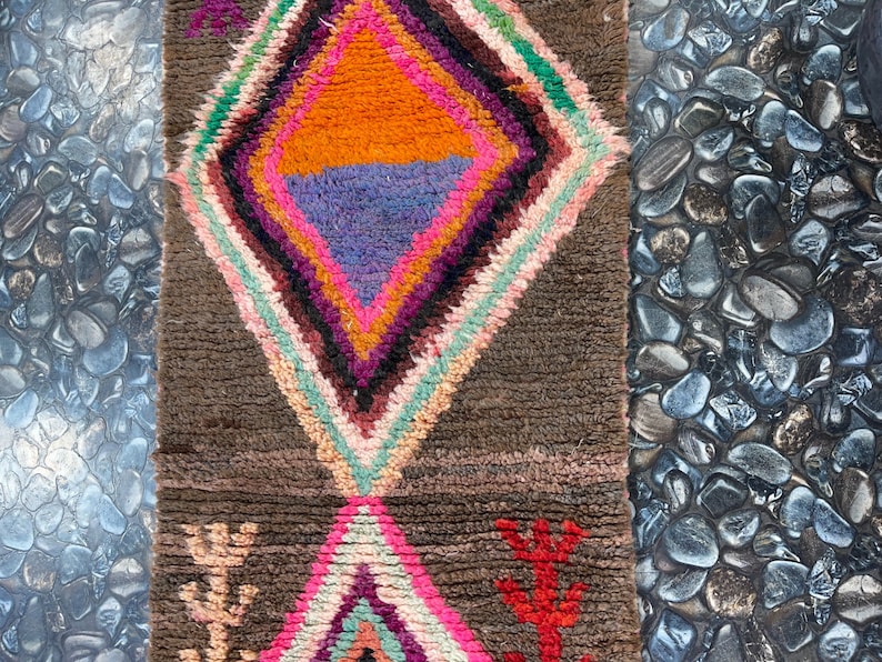 May include: A brown rug with a colorful diamond pattern in the center. The diamond is made up of pink, orange, purple, and green stripes. The rug has a fringe on the bottom.
