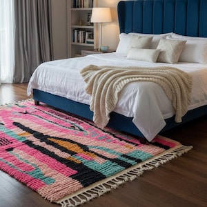 May include: A colorful, rectangular rug with a geometric pattern in shades of pink, black, teal, and orange. The rug is placed on a hardwood floor in front of a bed with a blue headboard. A white armchair and a bed with white linens are also visible.