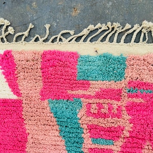 May include: Close-up of a colorful rug with a geometric pattern. The rug features a combination of bright pink, light pink, and teal squares and shapes. The top edge has a cream-colored border with knotted fringe.