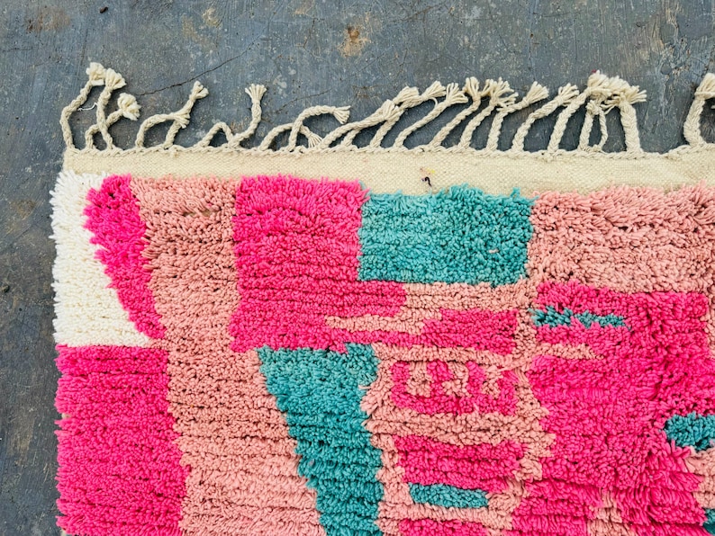 May include: Close-up of a colorful rug with a geometric pattern. The rug features a combination of bright pink, light pink, and teal squares and shapes. The top edge has a cream-colored border with knotted fringe.