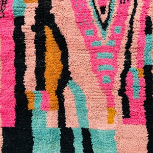 May include: Close-up of a colorful rug with a geometric pattern. The rug features a combination of pink, black, teal, and orange hues. The design includes vertical stripes and abstract shapes, creating a textured appearance.