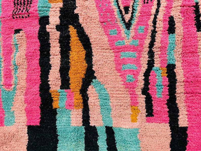 May include: Close-up of a colorful rug with a geometric pattern. The rug features a combination of pink, black, teal, and orange hues. The design includes vertical stripes and abstract shapes, creating a textured appearance.