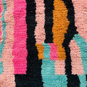 May include: Close-up of a colorful, textured rug with a geometric pattern. The rug features vertical stripes in vibrant pink, black, teal, peach, and gold. The plush texture and bold color scheme create a modern, eye-catching design.
