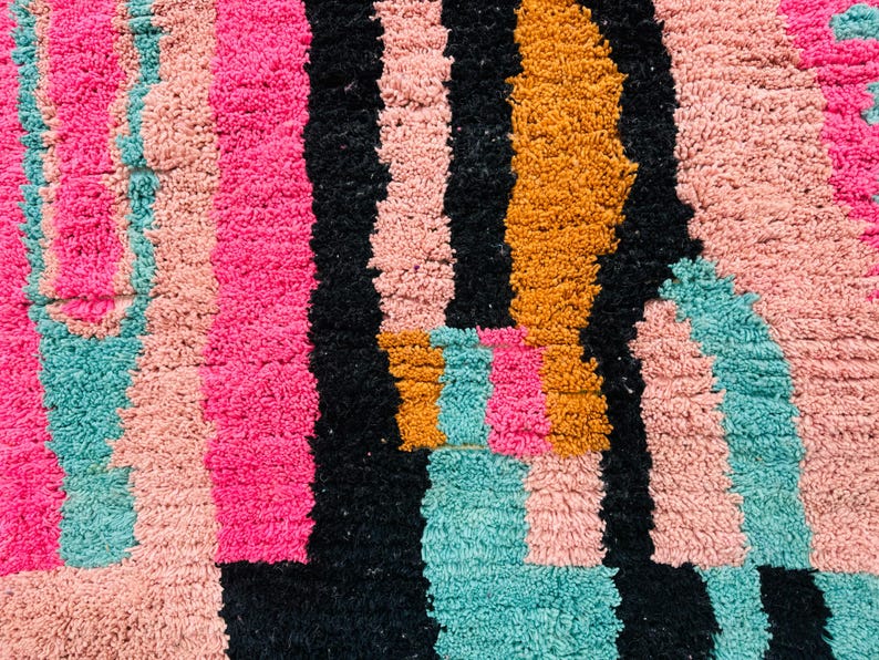 May include: Close-up of a colorful, textured rug with a geometric pattern. The rug features vertical stripes in vibrant pink, black, teal, peach, and gold. The plush texture and bold color scheme create a modern, eye-catching design.