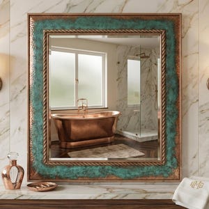 Op de afbeelding: Een rechthoekige wandspiegel met een frame van gehamerd koper, een rand met een groenachtige patina en een binnenlijst in de vorm van gedraaid touw. De spiegel reflecteert een badkamerinterieur met een koperen badkuip.
