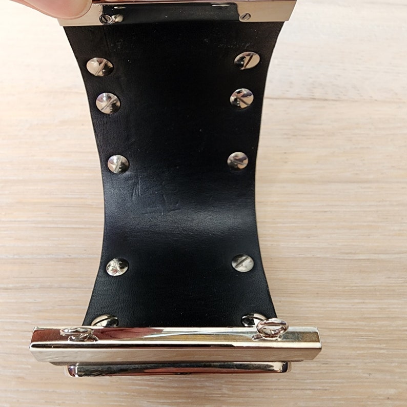 May include: A black leather cuff bracelet with silver metal accents and a silver metal clasp. The bracelet has a row of silver metal studs on each side.