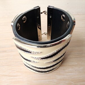 May include: A wide black leather cuff bracelet with a white and black striped design. The bracelet is decorated with clear rhinestones.