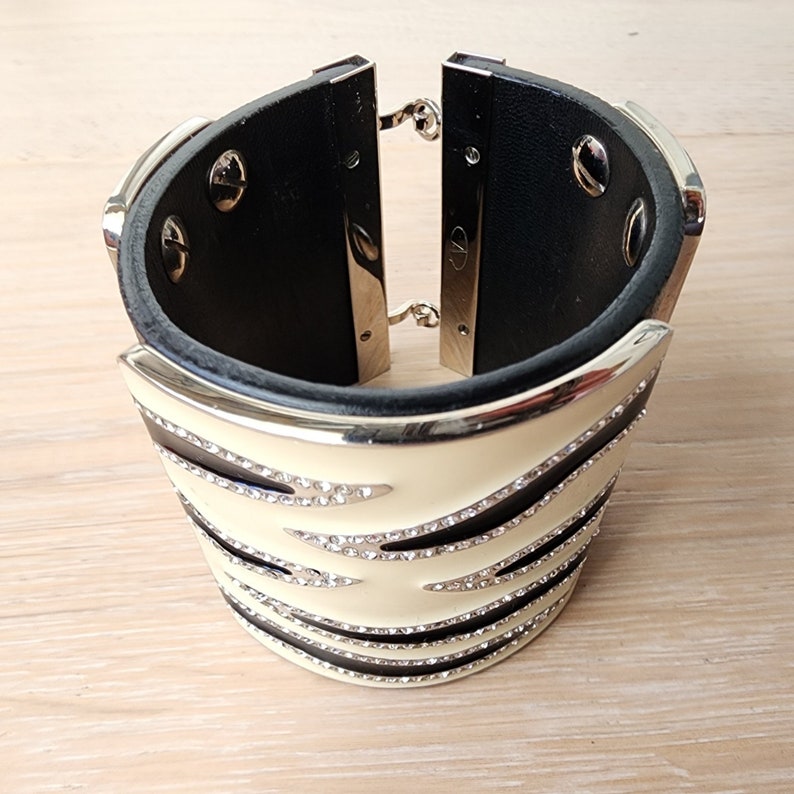 May include: A wide black leather cuff bracelet with a white and black striped design. The bracelet is decorated with clear rhinestones.