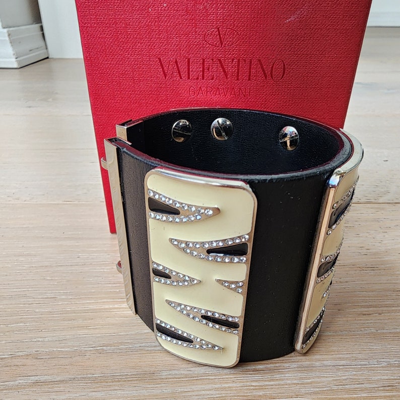May include: A black leather cuff bracelet with a silver metal trim and a white enamel design featuring a geometric pattern with rhinestones. The bracelet is accented with the text "VALENTINO GARAVANI".