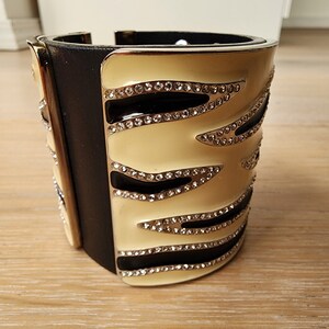 May include: A wide black leather cuff bracelet with a cream enamel design. The design features black lines and rows of clear rhinestones.
