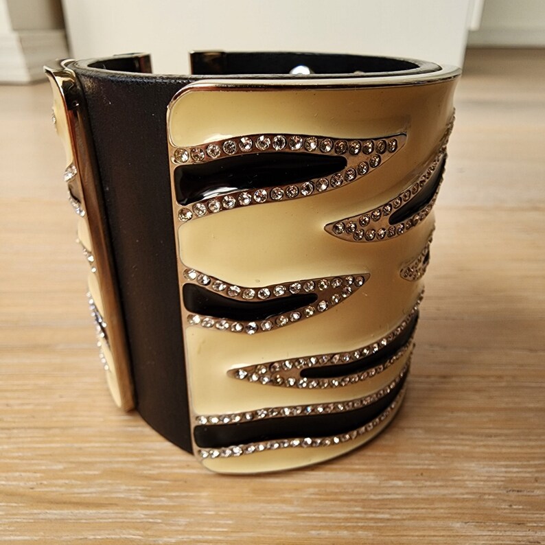 May include: A wide black leather cuff bracelet with a cream enamel design. The design features black lines and rows of clear rhinestones.