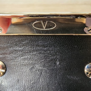 May include: Close-up of a black leather bag with a gold metal trim and a gold oval logo with the letter "V" inside.