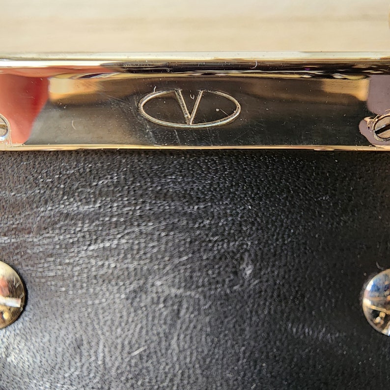 May include: Close-up of a black leather bag with a gold metal trim and a gold oval logo with the letter "V" inside.