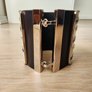 May include: A black leather cuff bracelet with silver metal accents and a decorative design. The bracelet has a hinged clasp.