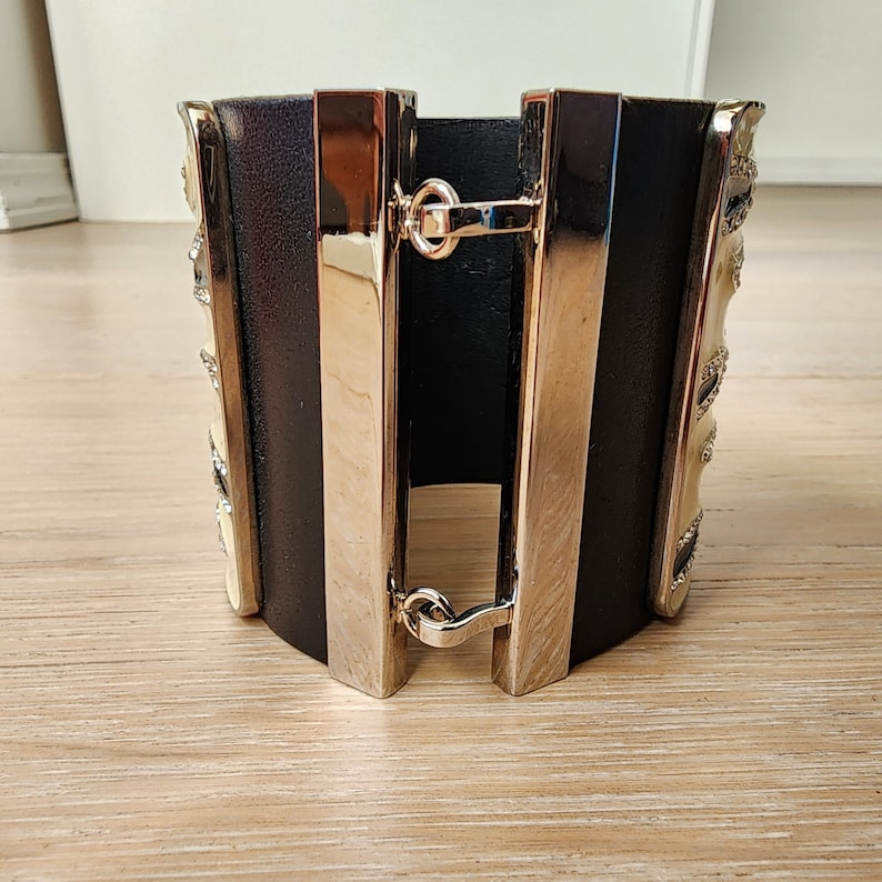 May include: A black leather cuff bracelet with silver metal accents and a decorative design. The bracelet has a hinged clasp.