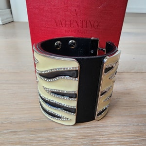 May include: A black and cream zebra-print cuff bracelet with crystal embellishments. The bracelet is made of leather and metal and features the Valentino Garavani logo.