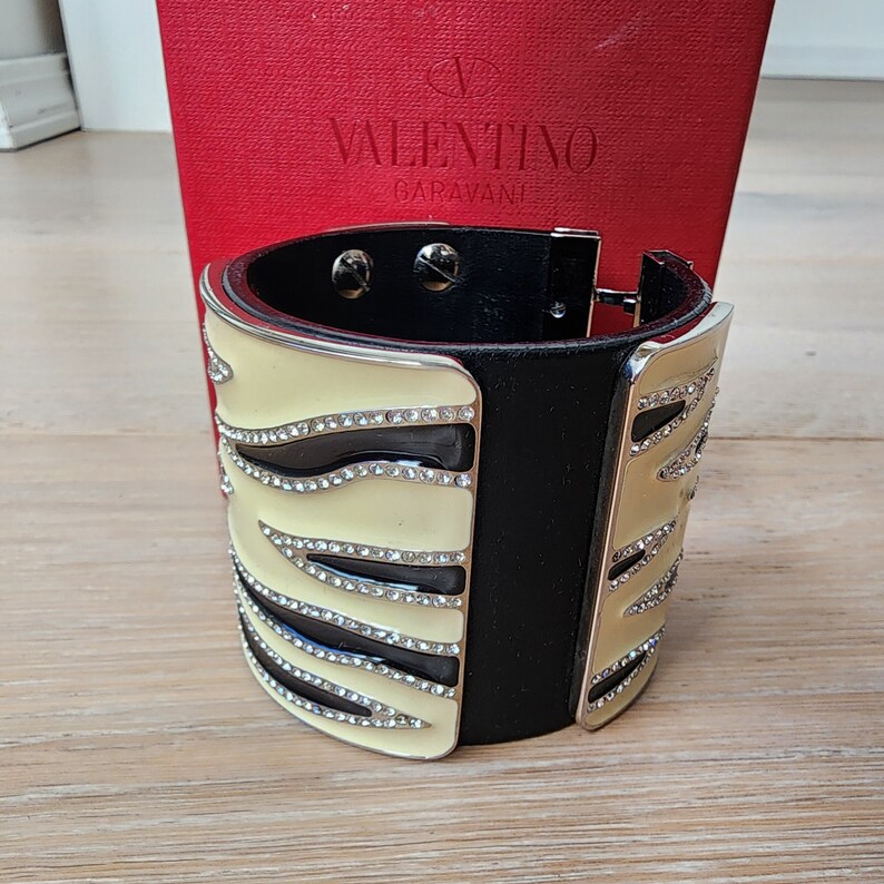 May include: A black and cream zebra-print cuff bracelet with crystal embellishments. The bracelet is made of leather and metal and features the Valentino Garavani logo.